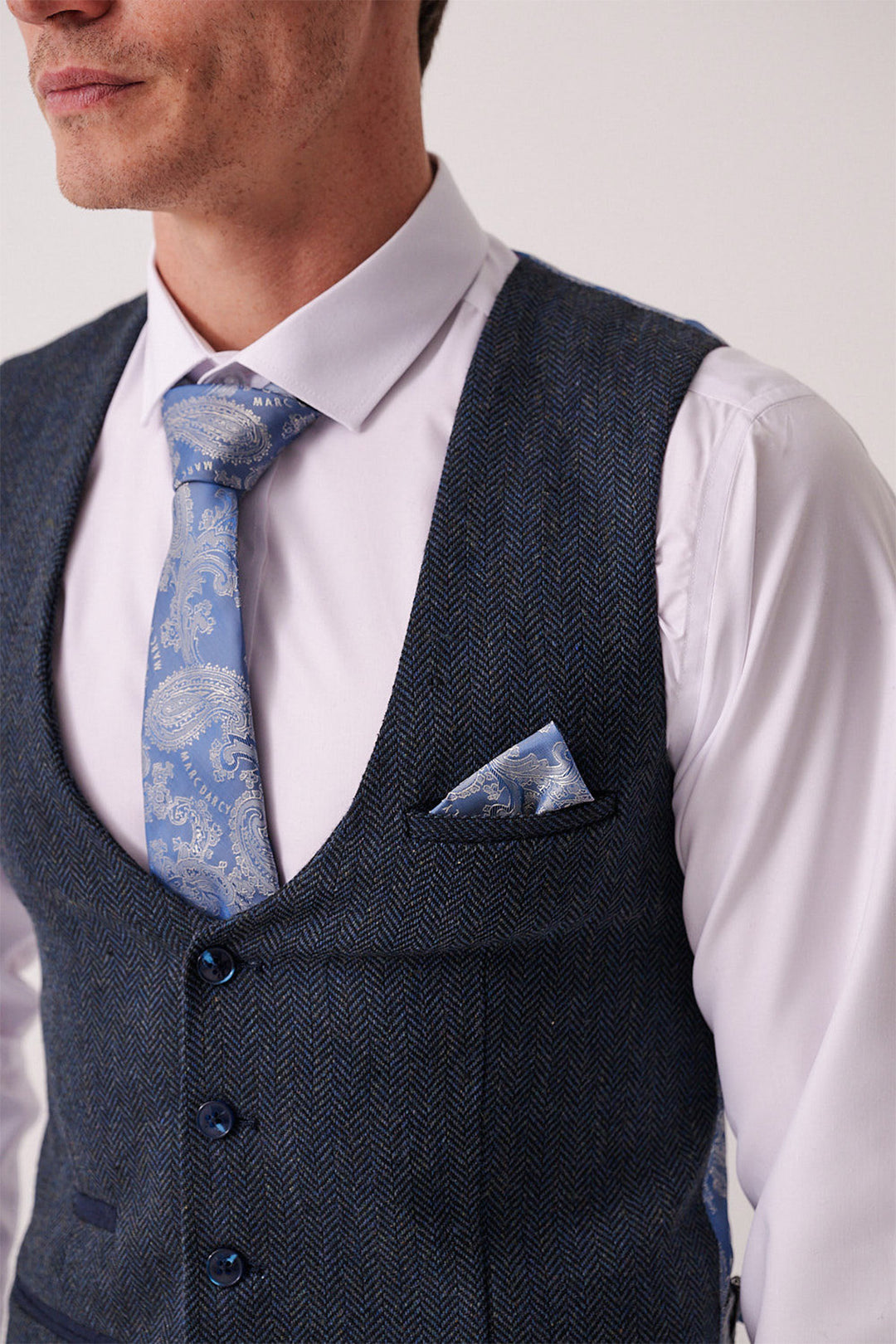 A man wearing the marcdarcy DEAN Blue Tweed Three Piece Suit, featuring a slim fit white shirt, blue herringbone vest, light blue paisley tie, and matching pocket square. The upper body is shown; his full face isn't visible.