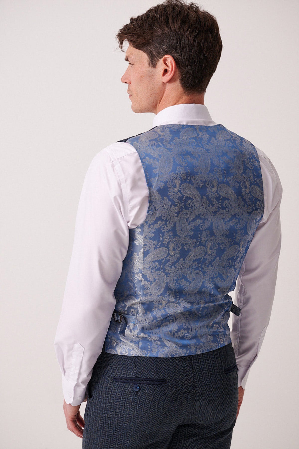 Back view of a man wearing a white dress shirt, dark herringbone pants, and the Marc Darcy DEAN Blue Tweed Waistcoat with paisley detailing.