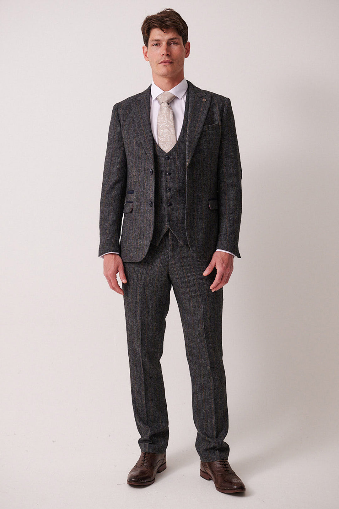 A man faces forward against a plain background, wearing the marcdarcy DOMINIC Blue Grey Tweed Three Piece Suit with a white dress shirt, light patterned tie, and brown dress shoes. His expression is neutral and posture relaxed.