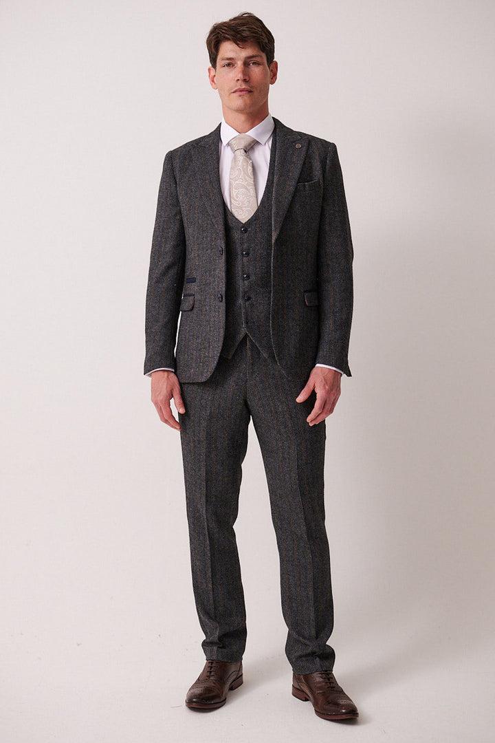 A man faces forward against a plain background, wearing the marcdarcy DOMINIC Blue Grey Tweed Three Piece Suit with a white dress shirt, light patterned tie, and brown dress shoes. His expression is neutral and posture relaxed.