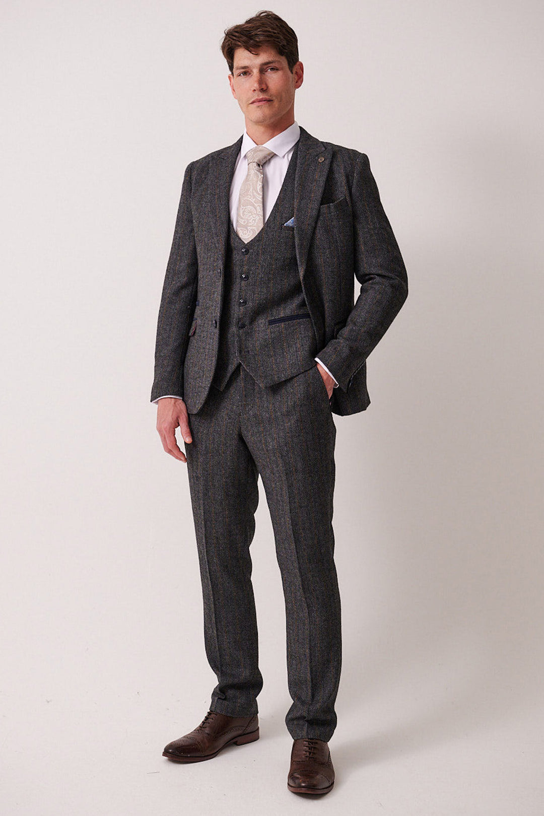 A man with short brown hair and a neutral expression stands against a plain light background, dressed in the marcdarcy DOMINIC Blue Grey Tweed Three Piece Suit, paired with a white shirt, patterned beige tie, and brown dress shoes.