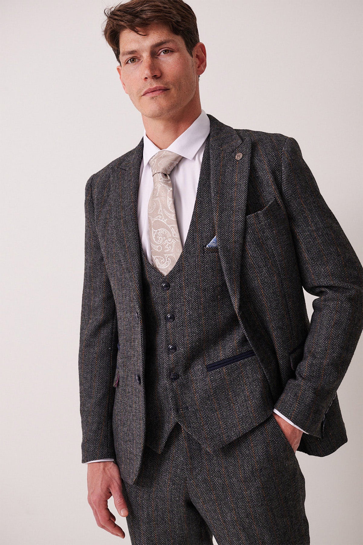 A man wears the marcdarcy DOMINIC Blue Grey Tweed Three Piece Suit with a white shirt and paisley tie, standing confidently against a plain light background with one hand in his pocket.