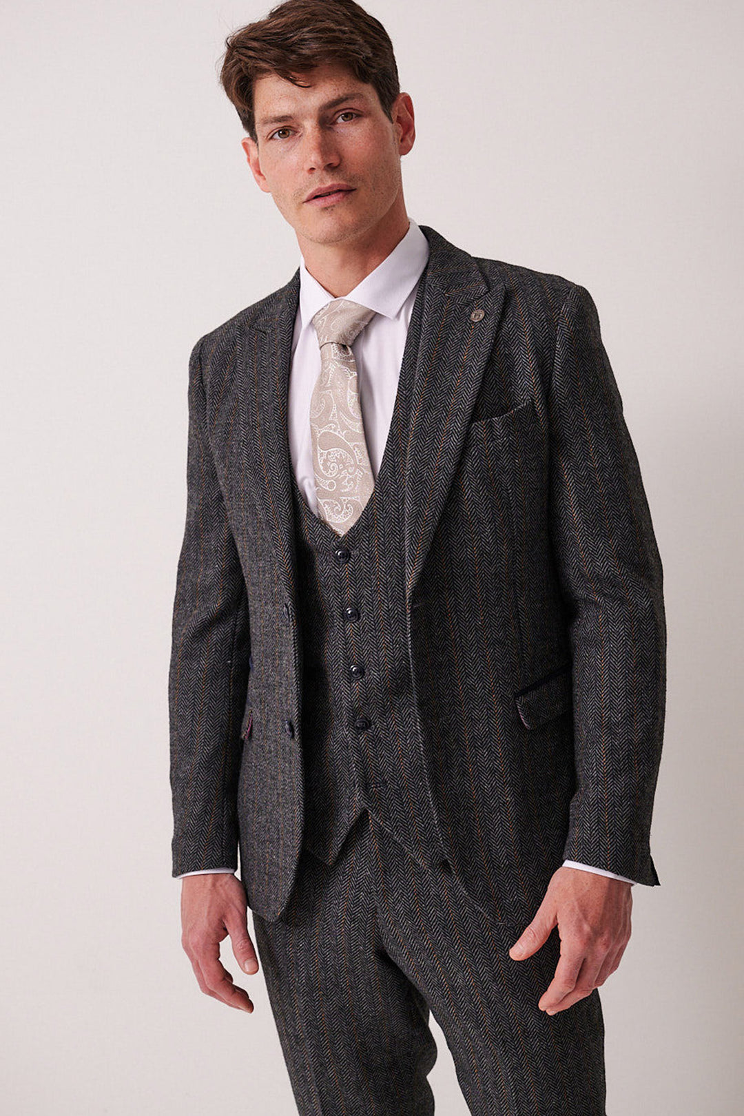 A man wearing the marcdarcy DOMINIC Blue Grey Tweed Blazer as part of a dark slim fit three-piece suit, paired with a white shirt and light paisley tie, stands against a plain light background, facing forward with a neutral expression.