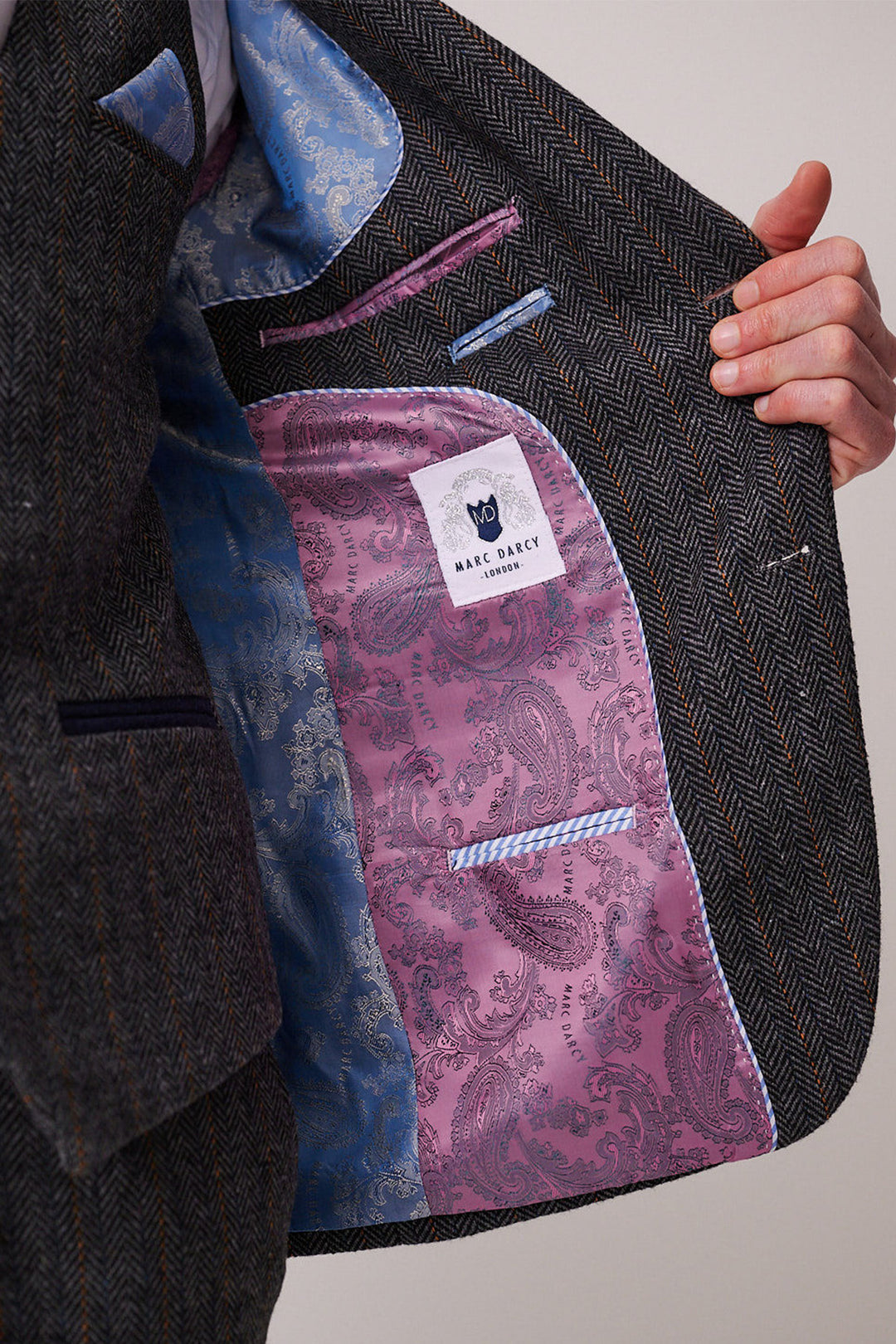 A person holds open a marcdarcy DOMINIC Blue Grey Tweed Three Piece Suit jacket to reveal a pink and blue paisley-patterned lining, with a visible marcdarcy label sewn inside.