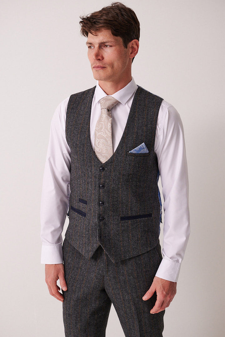 A man in a Marc Darcy suit with a DOMINIC Blue Grey Tweed Waistcoat, crisp white shirt, patterned tie, and blue pocket square stands against a plain background, looking slightly to the side.
