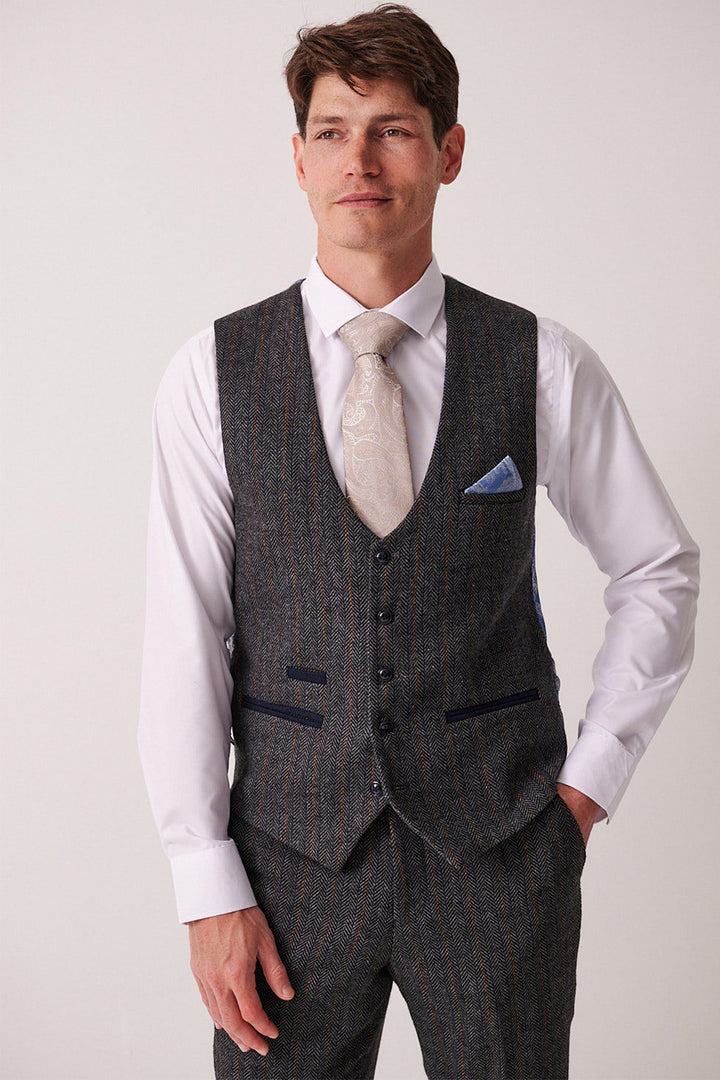 A man in the marcdarcy DOMINIC blue grey tweed three piece suit stands with one hand in his pocket, wearing a slim fit white shirt and patterned tie, gazing to the side with a gentle smile.