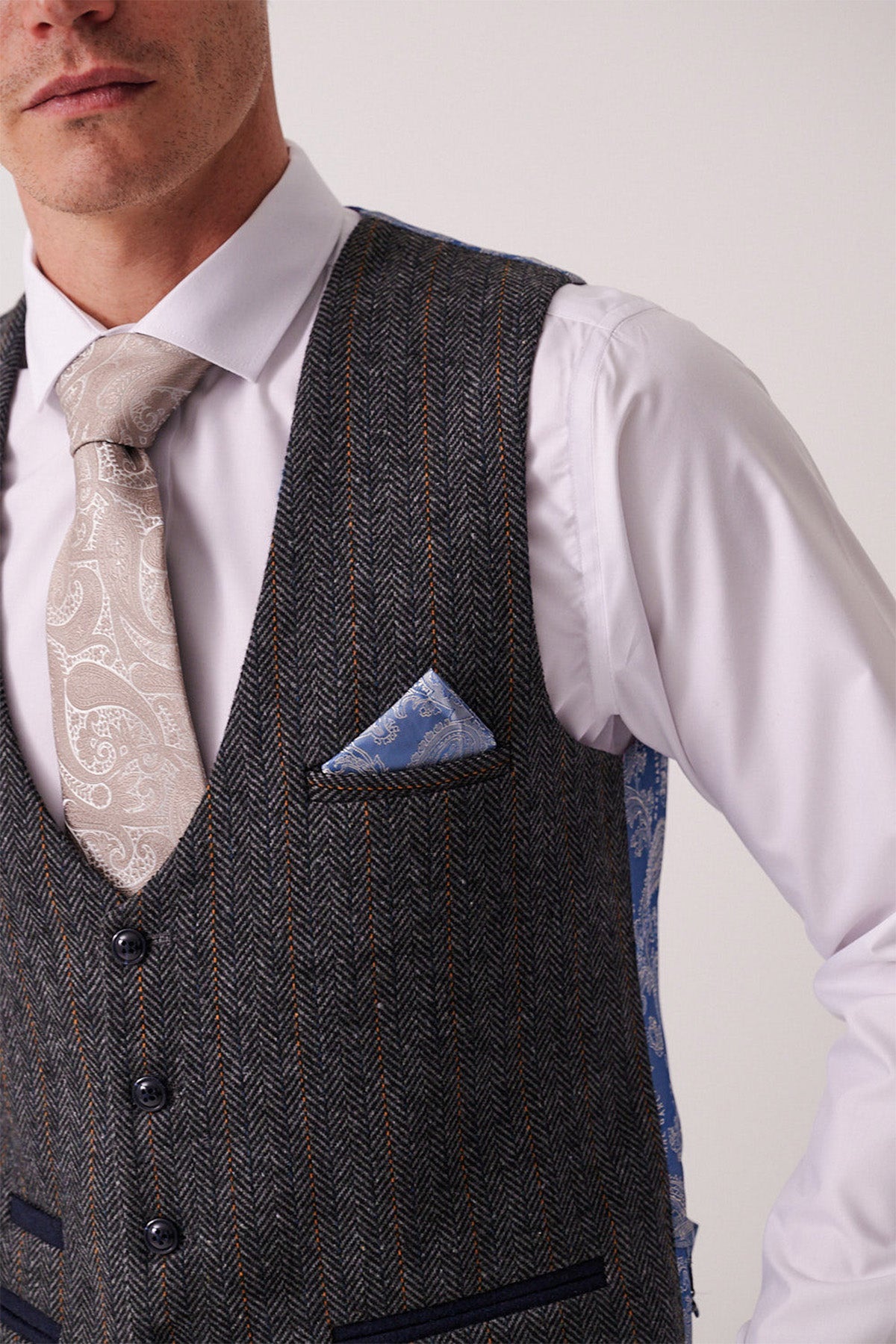 A man wears a white dress shirt, beige paisley tie, the Marc Darcy DOMINIC blue grey tweed waistcoat, and a blue patterned pocket square. The photo is cropped at the shoulders and chin, echoing classic Marc Darcy suit style.