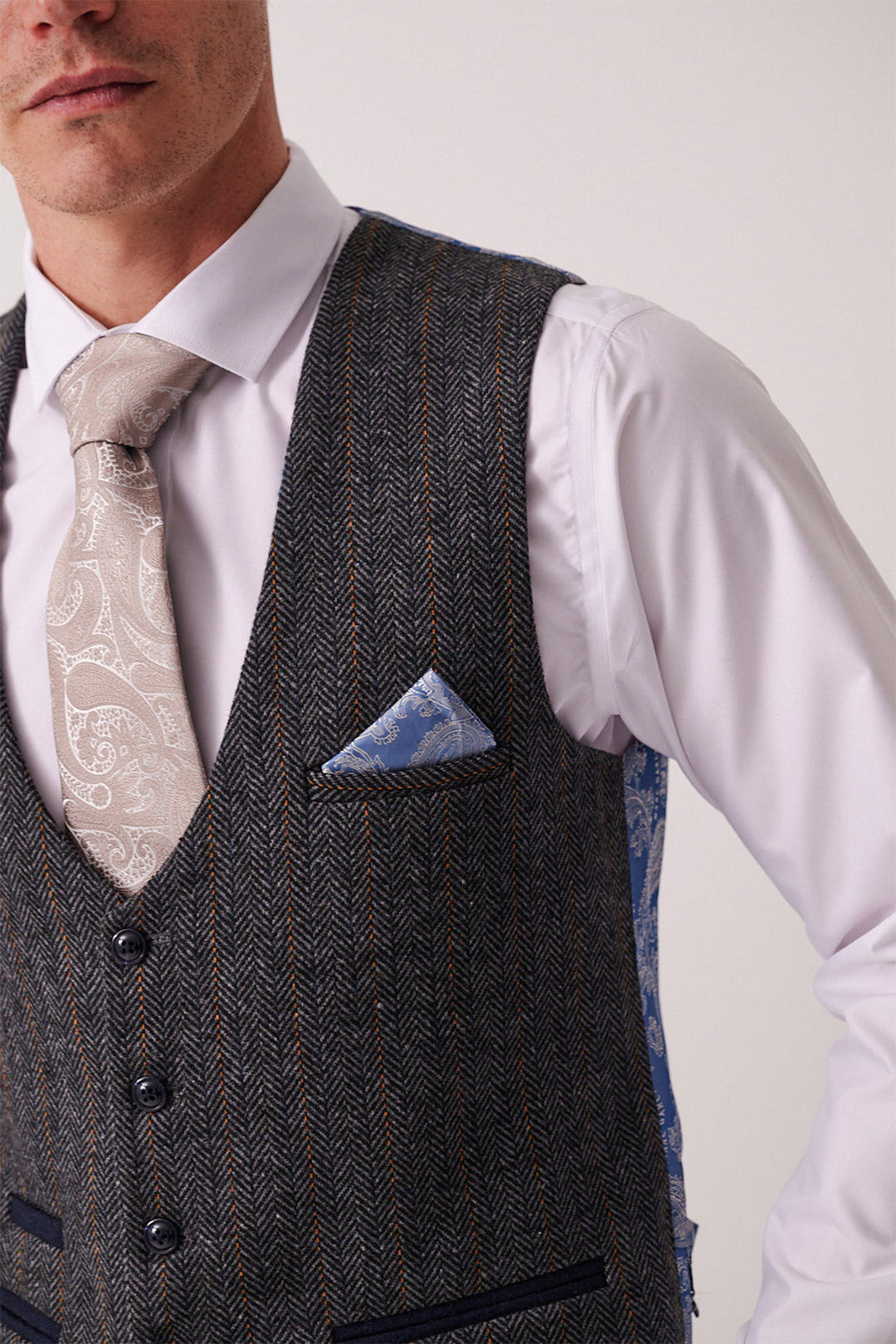 A man wears the marcdarcy DOMINIC Blue Grey Tweed Three Piece Suit styled with a white shirt, patterned beige tie, and blue pocket square. His slim-fit vest features gray herringbone with thin stripes. His face is partially out of frame.