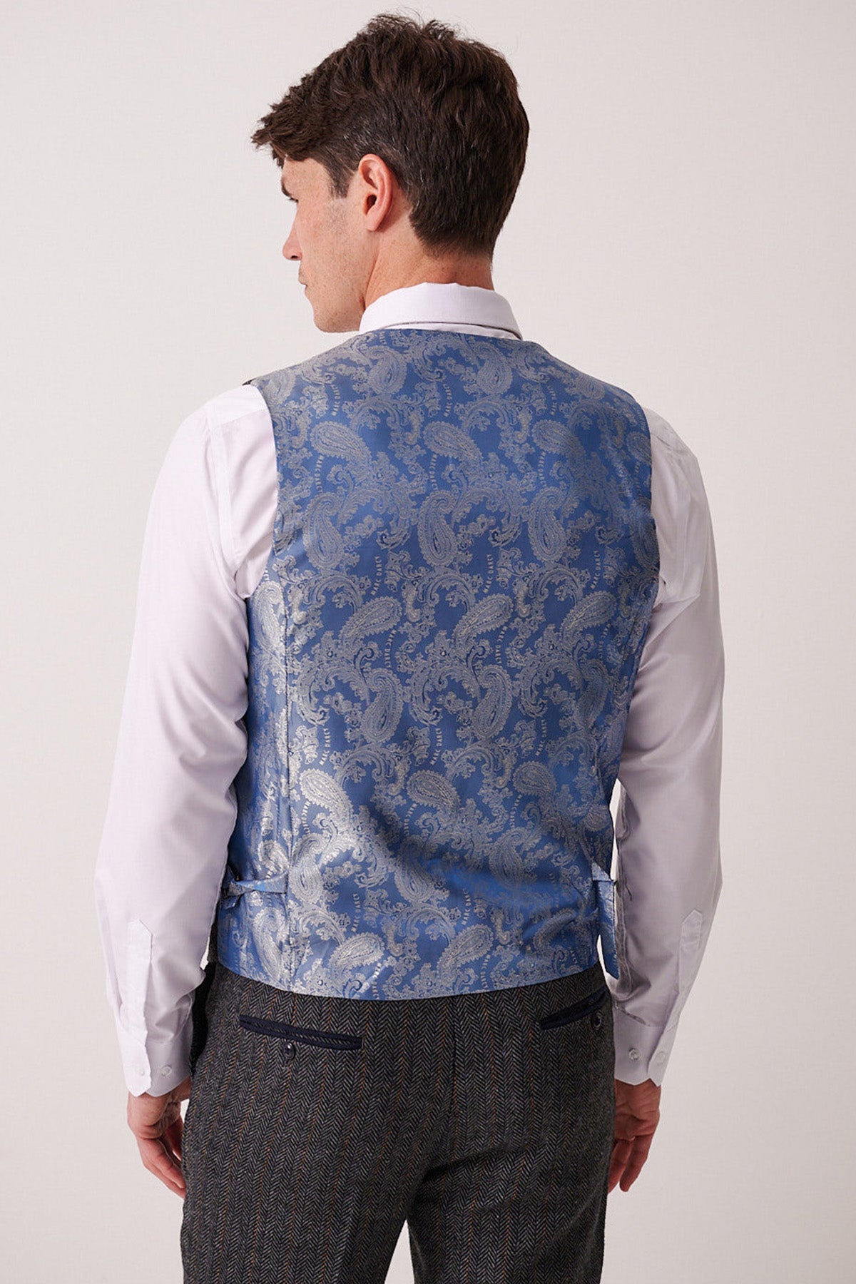 A man stands with his back to the camera, showcasing the detailed back of the Marc Darcy DOMINIC - Blue Grey Tweed Waistcoat, styled with a white dress shirt and gray Prince of Wales check trousers against a plain light background.