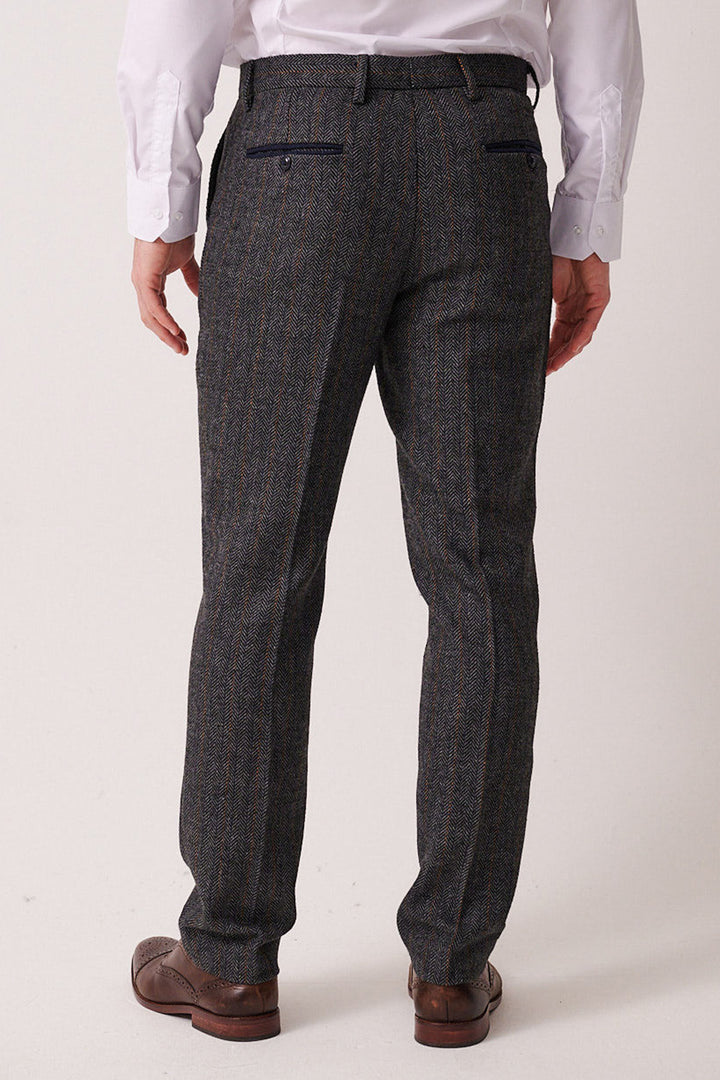 A man faces away, wearing the marcdarcy DOMINIC Blue Grey Tweed Three Piece Suit, featuring slim fit pants with two back pockets and brown dress shoes. The background is plain and light-colored.