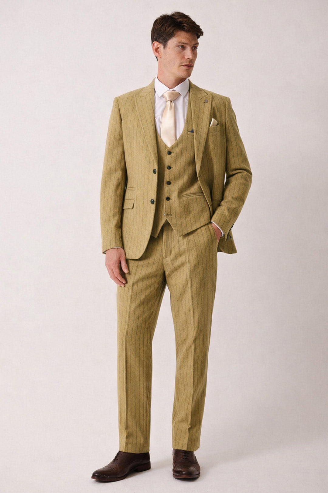 A man stands in the marcdarcy HANSON Moss Tweed Pinstripe Three Piece Suit with a white shirt, cream tie, and brown dress shoes. One hand in his pocket, he looks slightly to the side against a plain background.