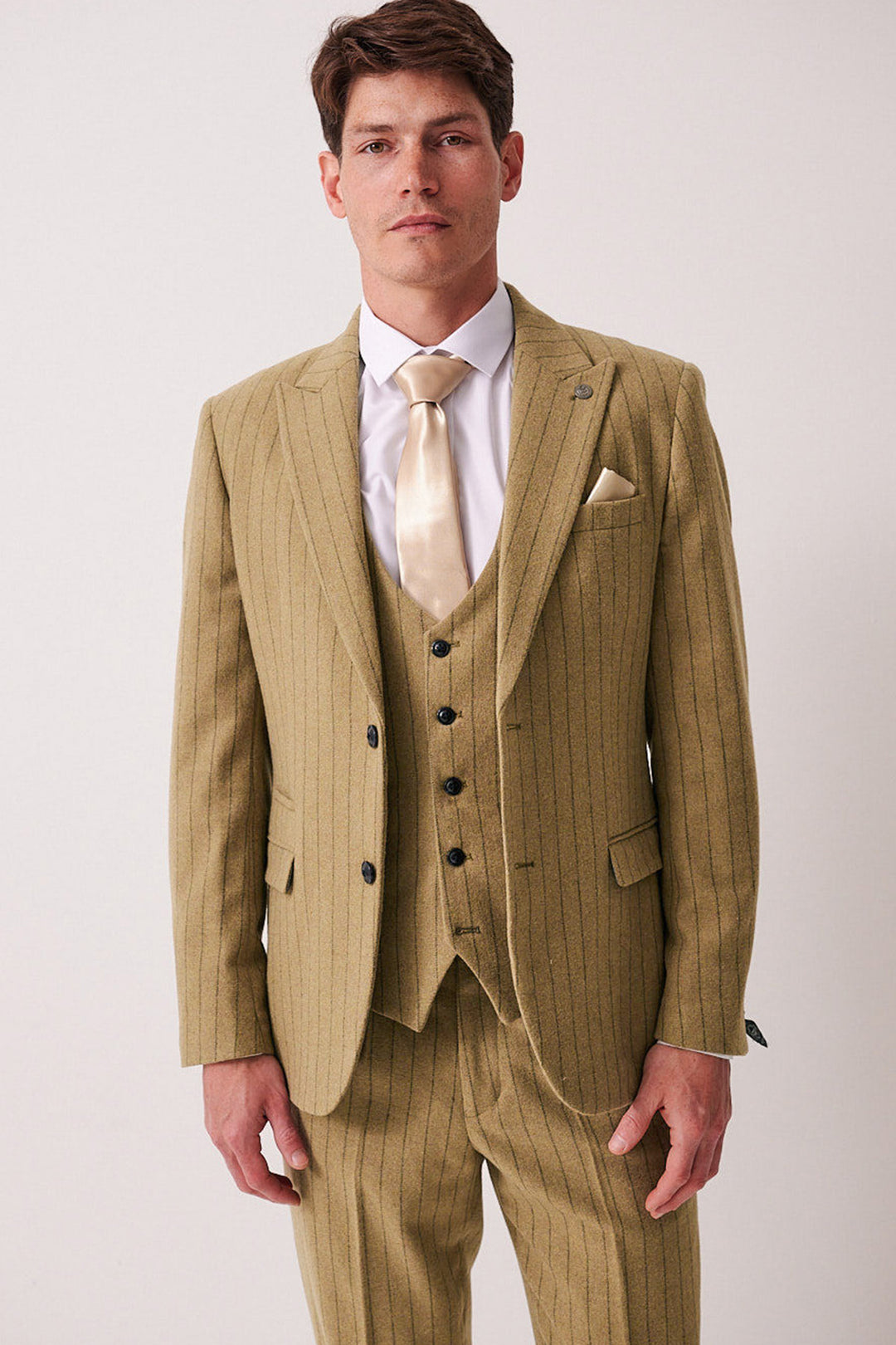 A man stands facing forward in the marcdarcy HANSON Moss Tweed Pinstripe Three Piece Suit with a white dress shirt and beige tie. He has short brown hair, a neutral expression, and is posed against a plain light background.