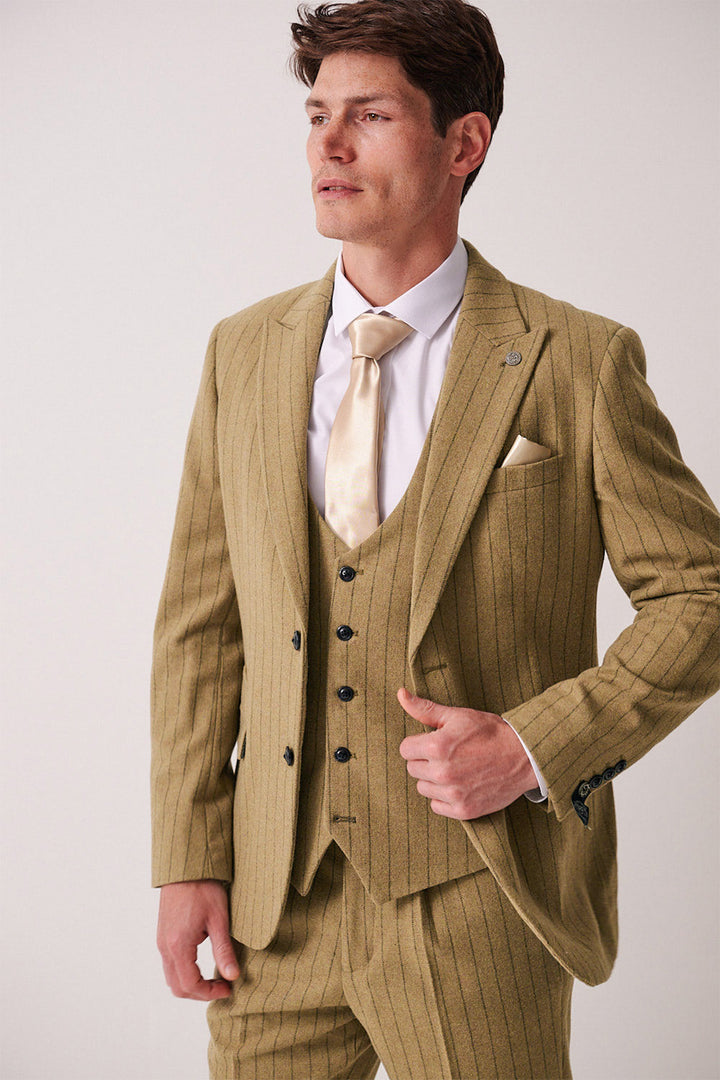 A man in a marcdarcy HANSON Moss Tweed Pinstripe Blazer with matching vest, white shirt, cream tie, and pocket square stands against a plain light background, looking aside while adjusting his jacket.