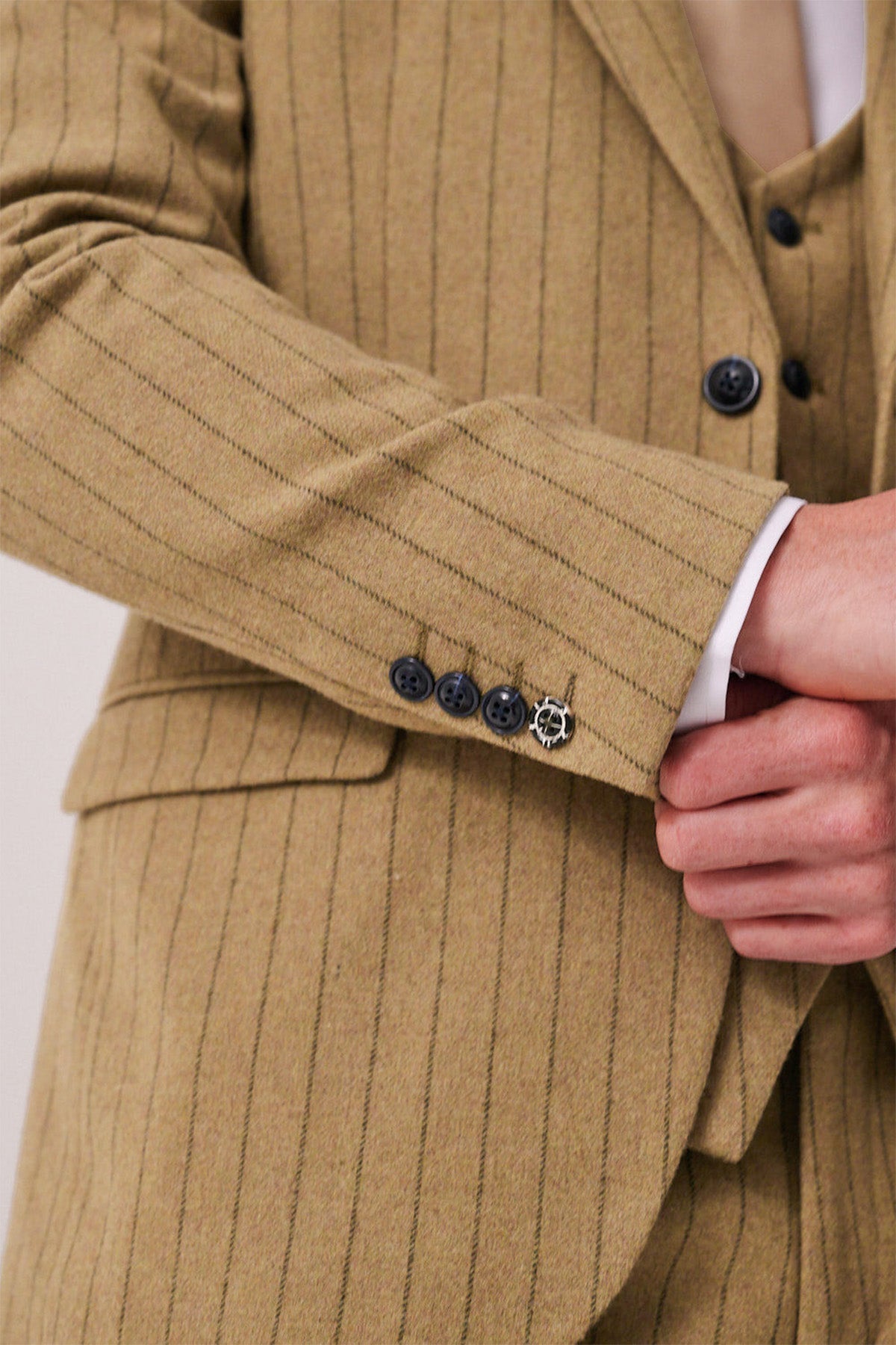 A close-up of a person adjusting the HANSON - Moss Tweed Pinstripe Blazer by marcdarcy, featuring tan fabric with pinstripes and four dark cuff buttons, one adorned with a small decorative metal accent.