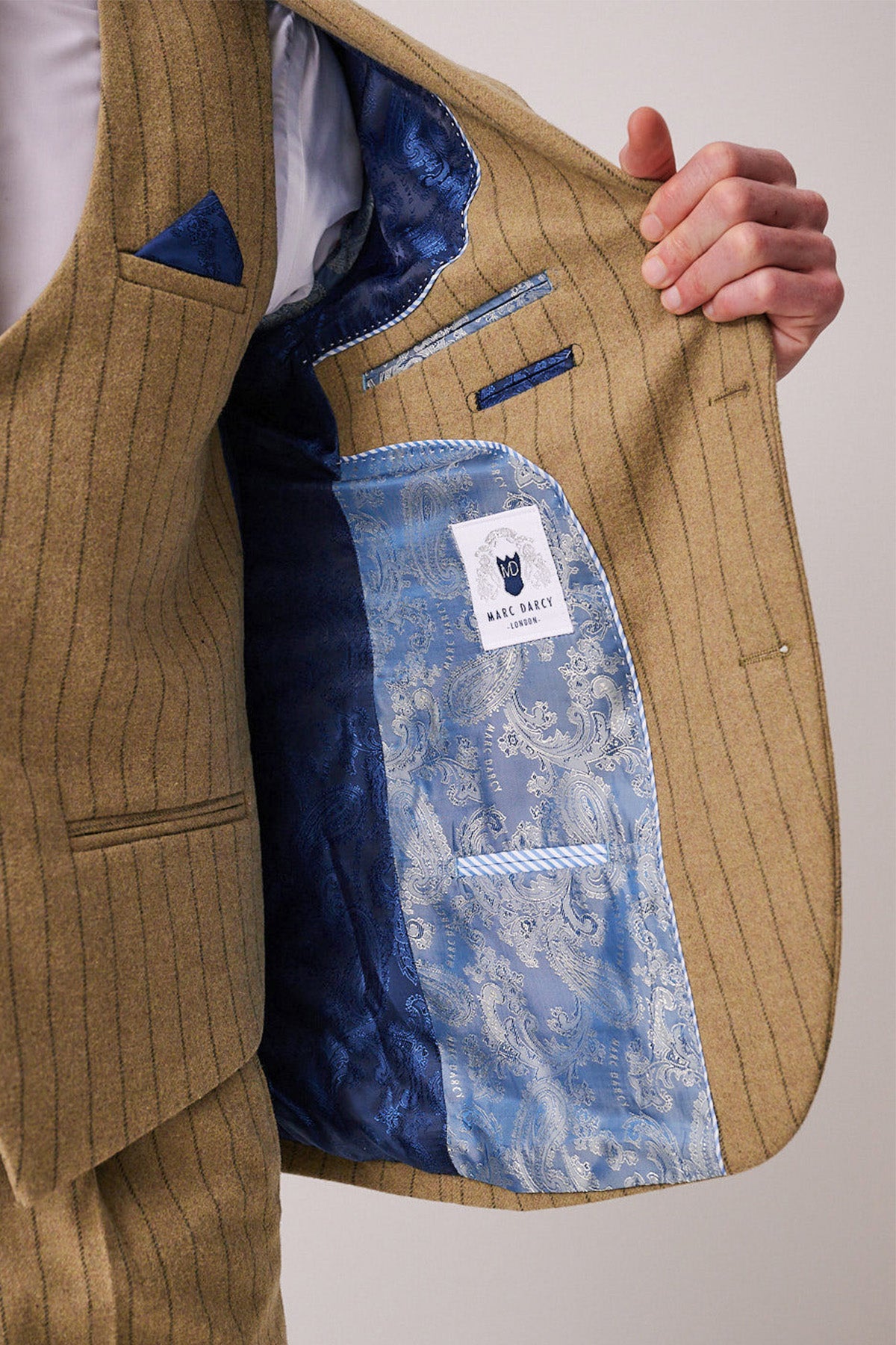 A person holds open a tan pinstripe HANSON - Moss Tweed Pinstripe Blazer by marcdarcy, revealing a blue paisley-patterned lining, a blue pocket square, and marcdarcy label—highlighting the brand’s attention to detail.
