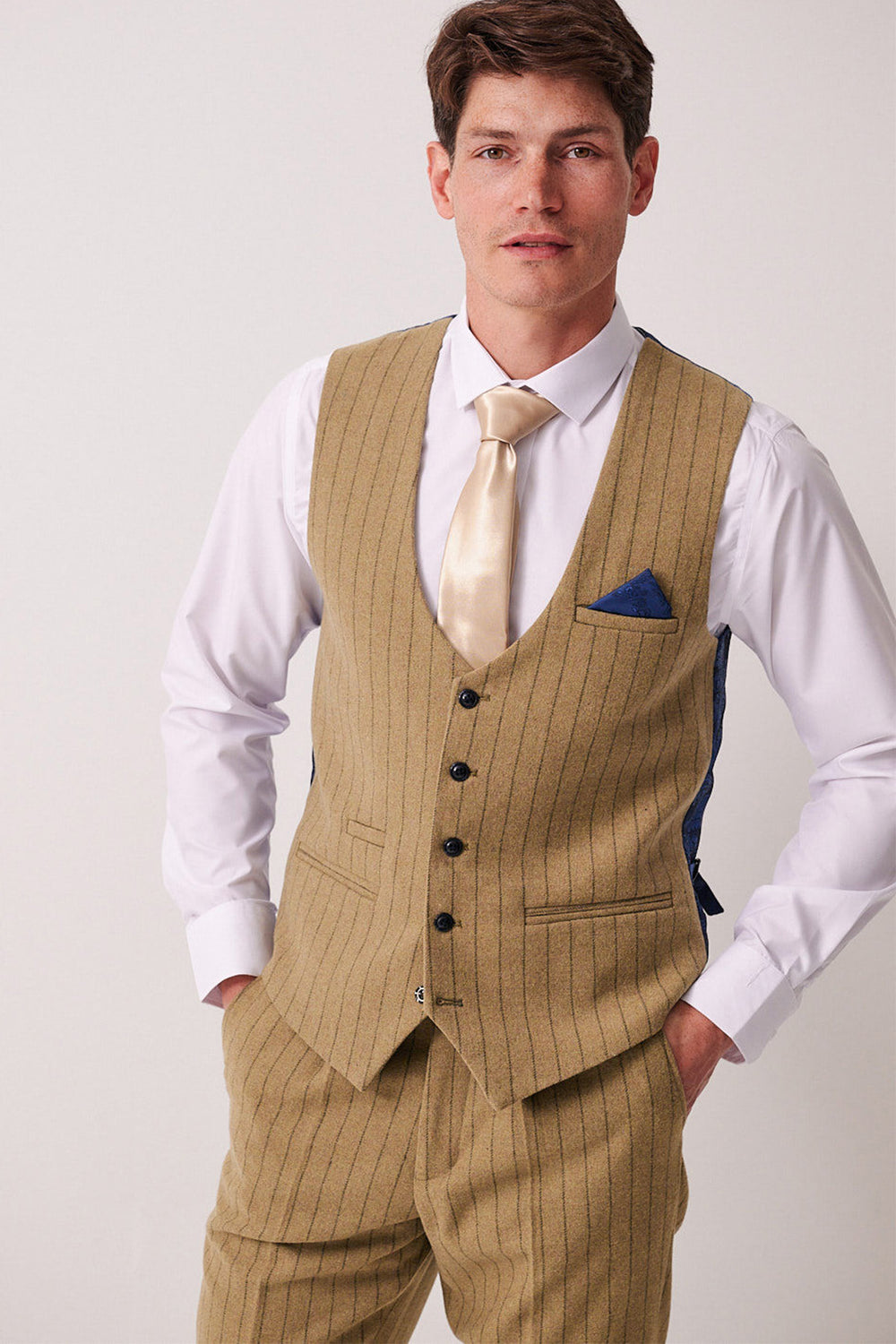 A man wearing the Marc Darcy HANSON Moss Tweed Pinstripe Waistcoat with matching trousers, a white shirt, gold satin tie, and blue pocket square stands facing the camera with his hands in his pockets.