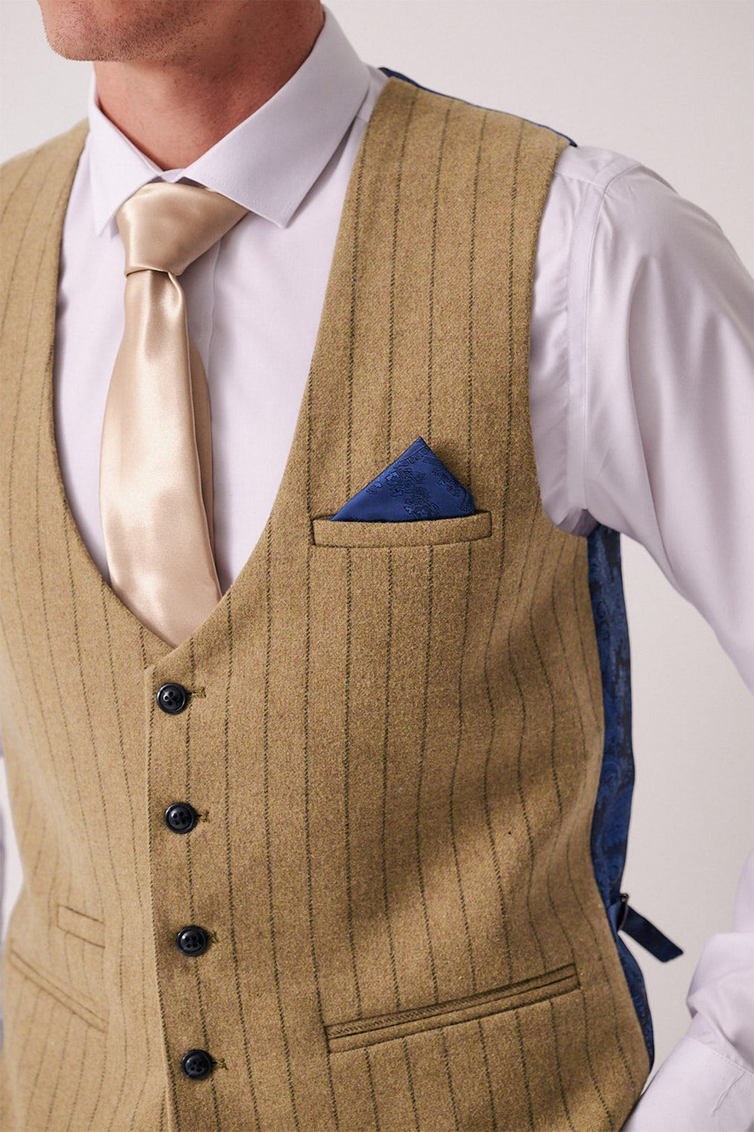 A man wears the Marc Darcy HANSON Moss Tweed Pinstripe Waistcoat over a white dress shirt, paired with a beige satin tie and a blue patterned pocket square in the breast pocket.