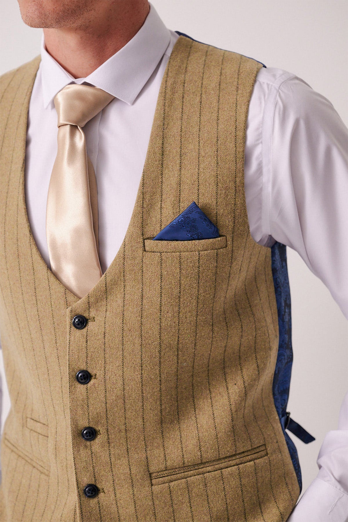 A man wears the Marc Darcy HANSON Moss Tweed Pinstripe Waistcoat over a white dress shirt, paired with a beige satin tie and a blue patterned pocket square in the breast pocket.