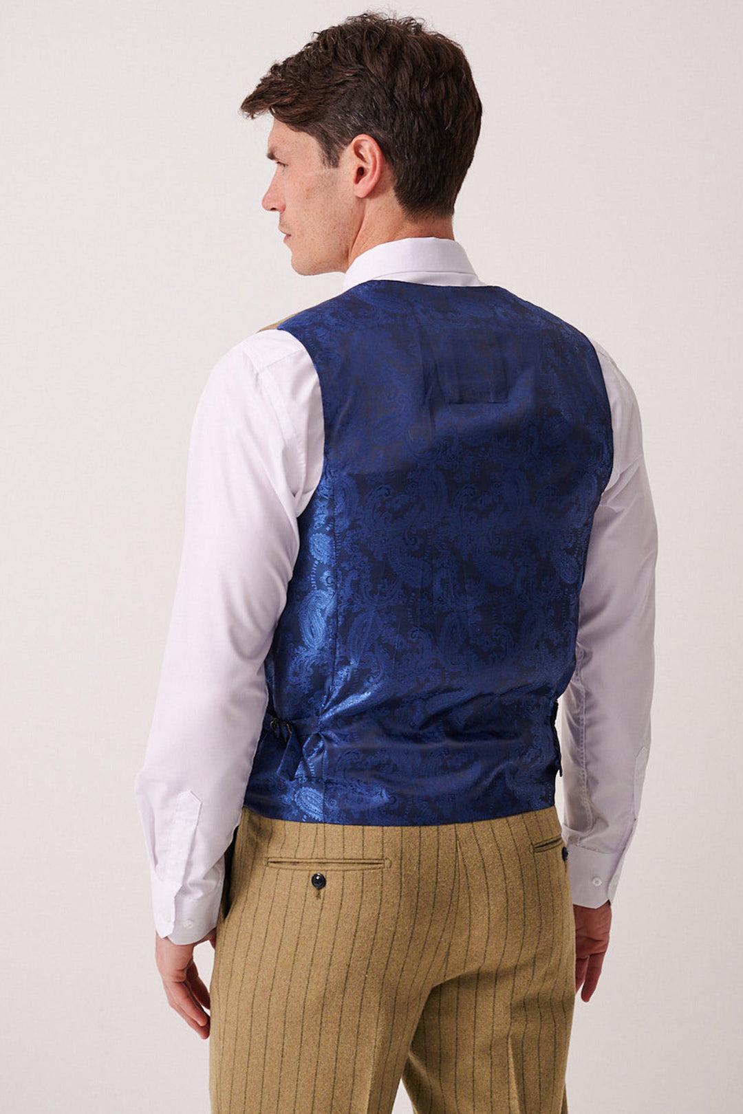 A man is seen from the back wearing a white shirt, tan pinstripe trousers, and the Marc Darcy HANSON - Moss Tweed Pinstripe Waistcoat, standing against a plain light background.