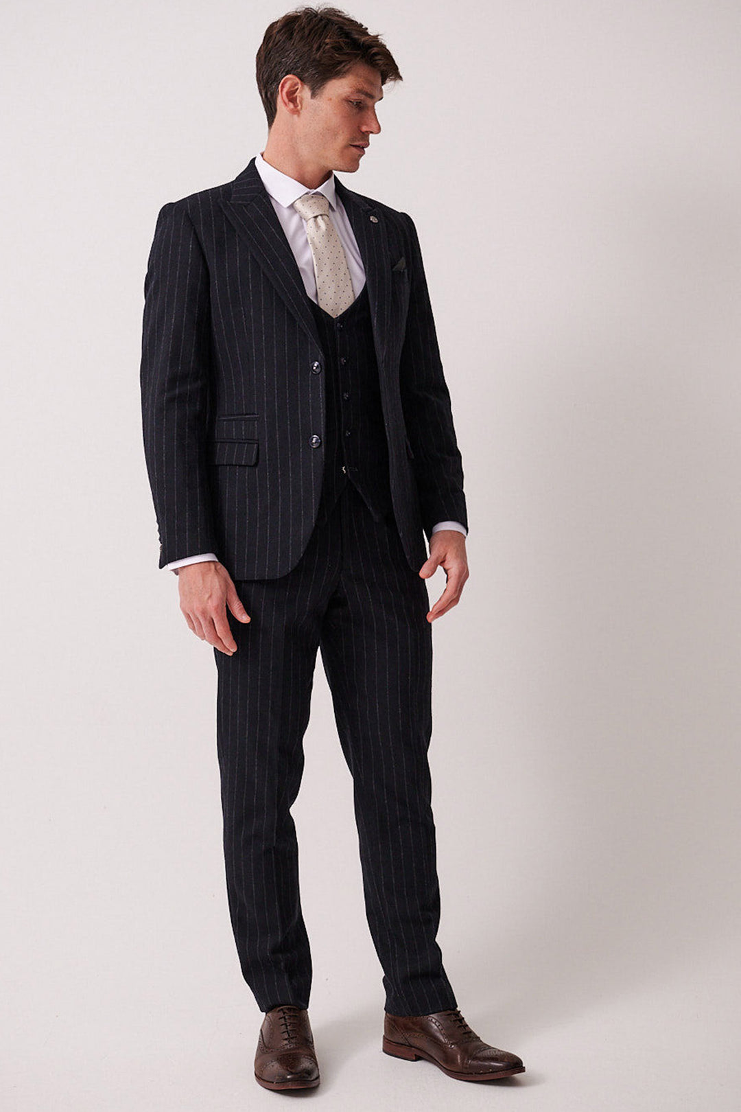 A man wears the marcdarcy HANSON Navy Tweed Pinstripe Three Piece Suit with a white dress shirt, cream tie, and brown shoes. Against a plain background, he looks down to his left with hands relaxed by his sides.