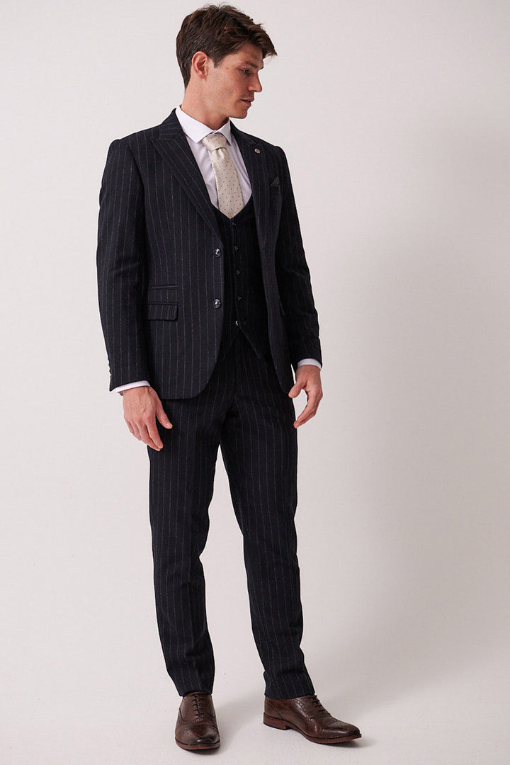 A man wears the marcdarcy HANSON Navy Tweed Pinstripe Three Piece Suit with a white dress shirt, cream tie, and brown shoes. Against a plain background, he looks down to his left with hands relaxed by his sides.