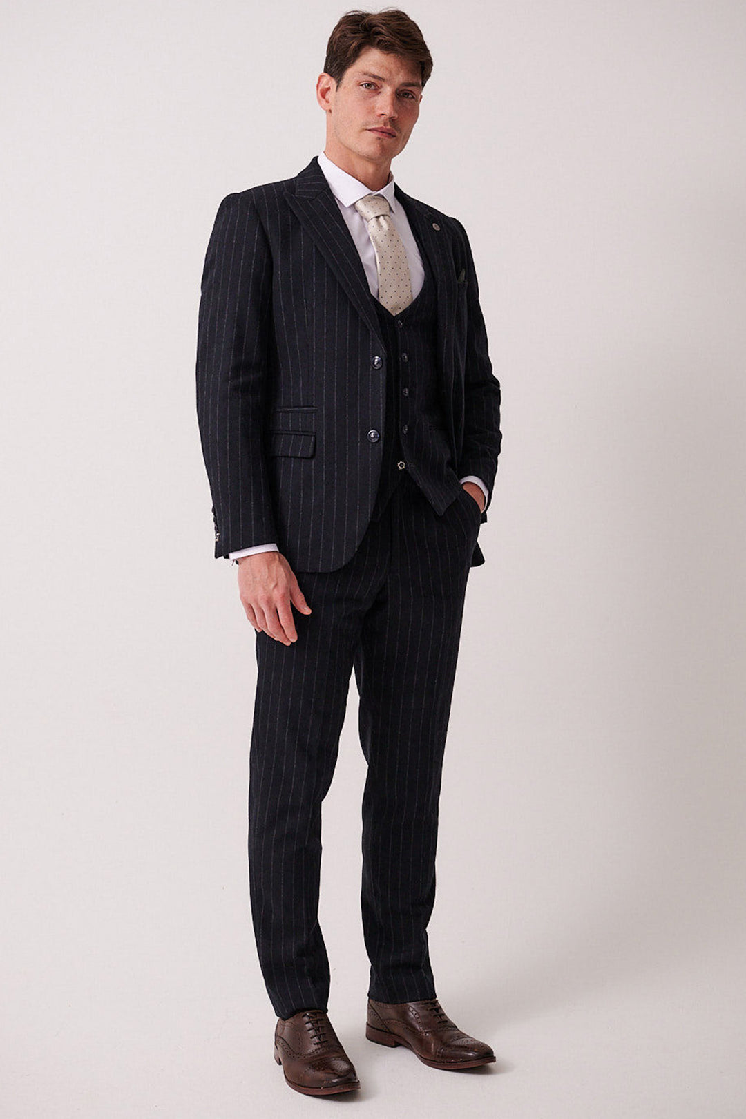 A man wears the marcdarcy HANSON Navy Tweed Pinstripe Three Piece Suit with a white shirt, light tie, and brown dress shoes, standing against a plain light background and looking at the camera with one hand in his pocket.