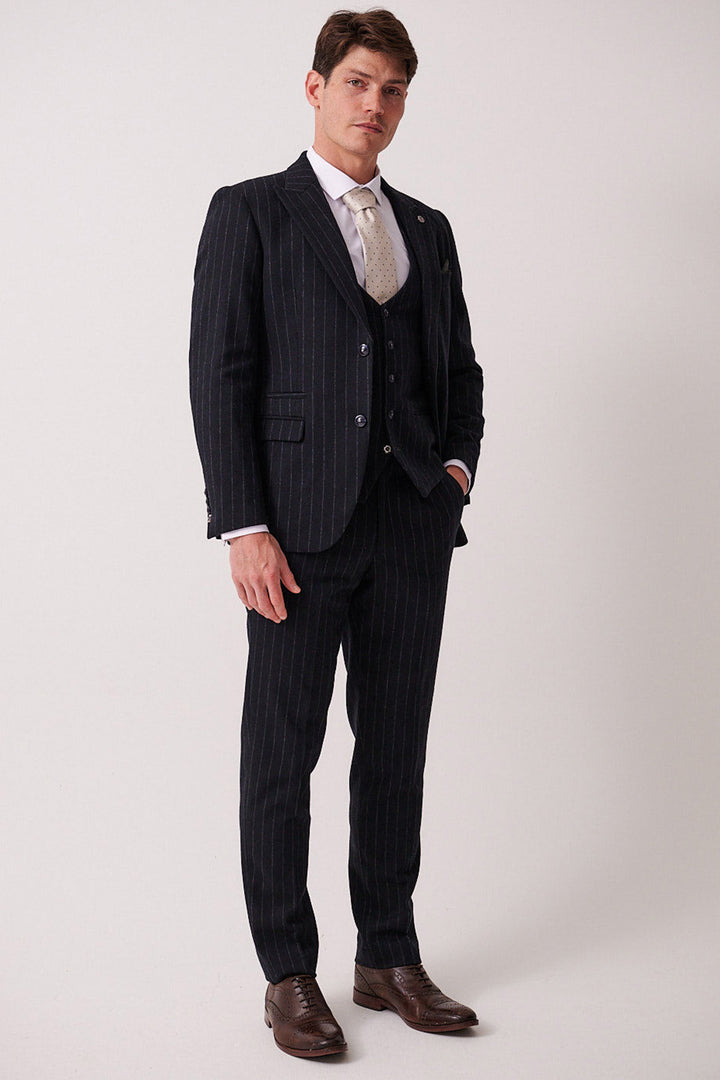 A man wears the marcdarcy HANSON Navy Tweed Pinstripe Three Piece Suit with a white shirt, light tie, and brown dress shoes, standing against a plain light background and looking at the camera with one hand in his pocket.