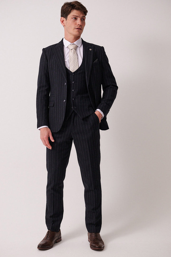 A man wears the marcdarcy HANSON Navy Tweed Pinstripe Three Piece Suit with a white dress shirt, light patterned tie, and brown dress shoes, standing against a plain background with one hand in his pocket and looking to the side.