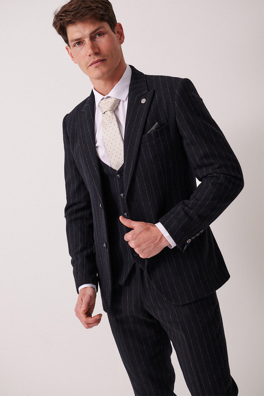 A man in a marcdarcy HANSON navy tweed pinstripe three piece suit, paired with a white dress shirt and light polka-dot tie, stands against a plain light background, confidently looking at the camera while adjusting his jacket.