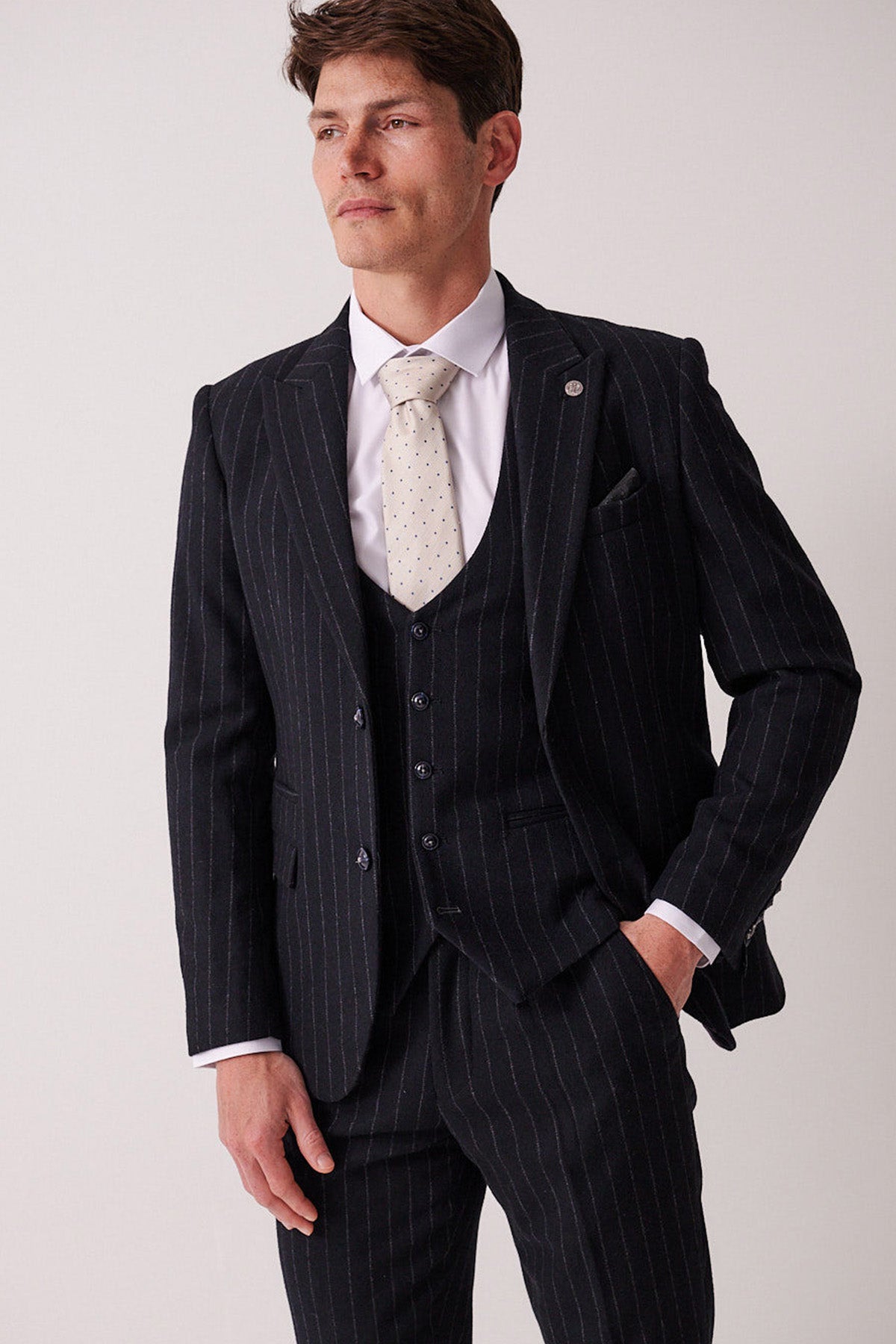 A man wearing the marcdarcy HANSON Navy Tweed Pinstripe Three Piece Suit with a white shirt and light polka dot tie stands with one hand in his pocket, gazing off to the side against a plain background.