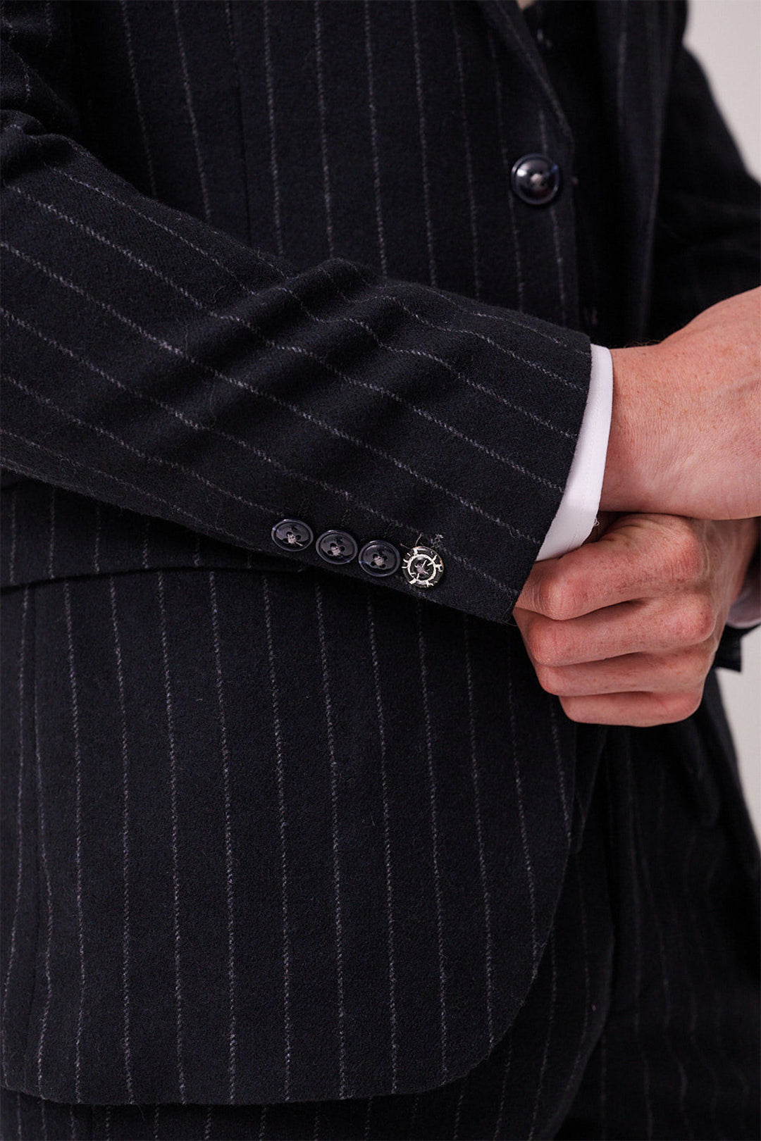 A person in a HANSON - Navy Tweed Pinstripe Three Piece Suit by marcdarcy adjusts their sleeve, revealing a silver ship’s wheel cufflink near the jacket cuff.