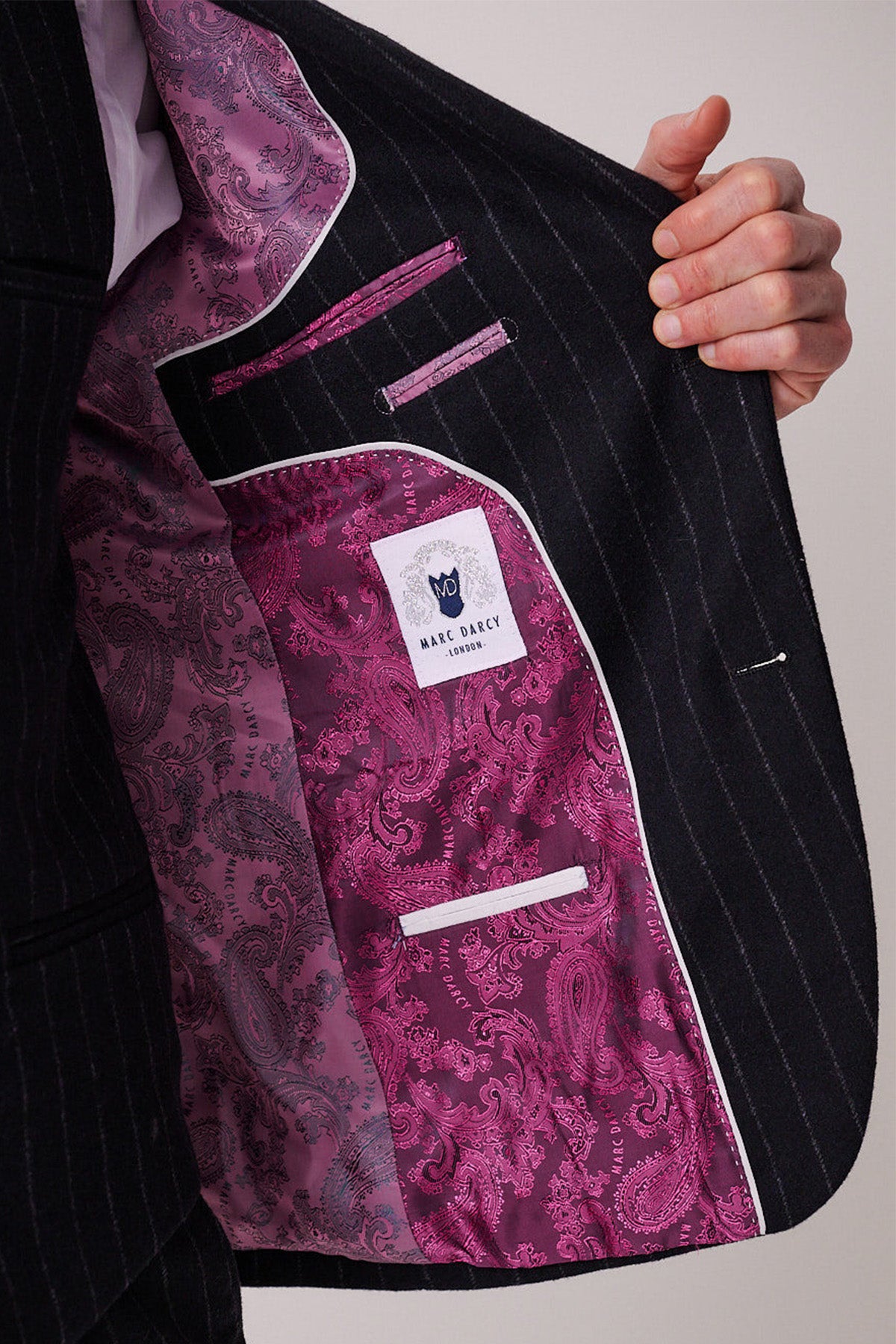 A person holds open a HANSON navy tweed pinstripe three piece suit jacket by marcdarcy, revealing a vibrant pink paisley-patterned lining and a white “marcdarcy” label.