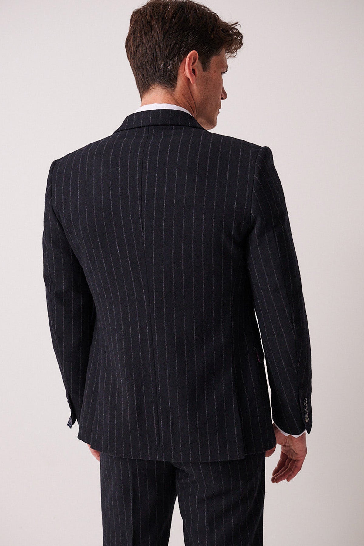A man with brown hair faces away, wearing the marcdarcy HANSON Navy Tweed Pinstripe Blazer and matching trousers. He stands against a plain light background, highlighting the sharp cut of his suit.