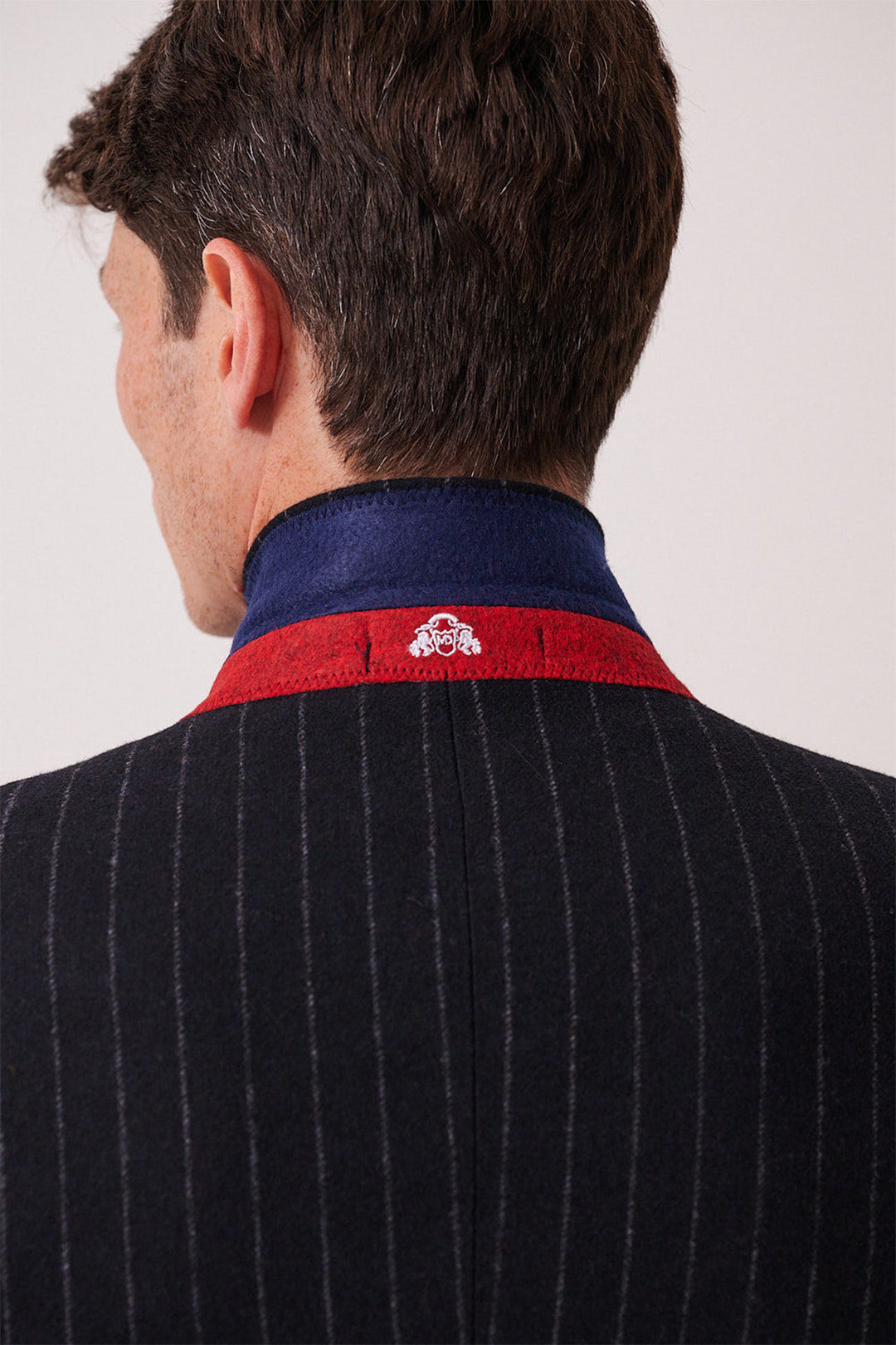 A man is shown from behind wearing the marcdarcy HANSON navy tweed pinstripe three piece suit. The collar is turned up, revealing a blue lining with red trim and a small white embroidered symbol at its center.