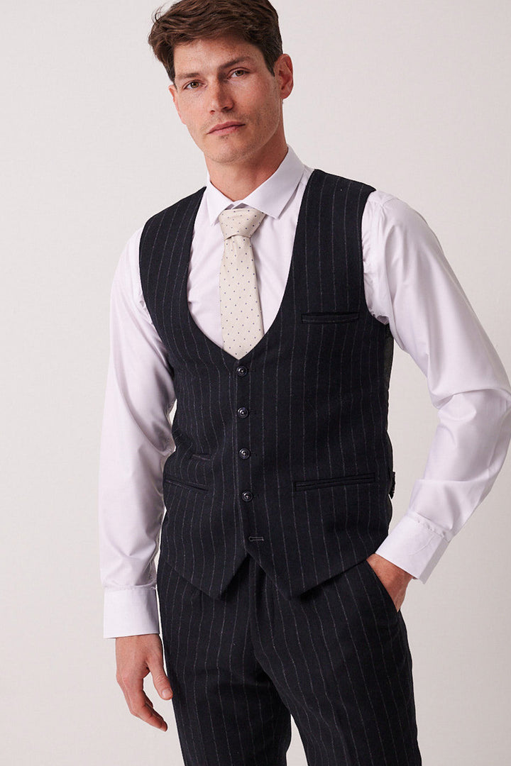 A man in the marcdarcy HANSON navy tweed pinstripe three piece suit stands facing forward, wearing a slim fit vest and pants with a white dress shirt and a light-colored polka dot tie, showing a neutral expression.