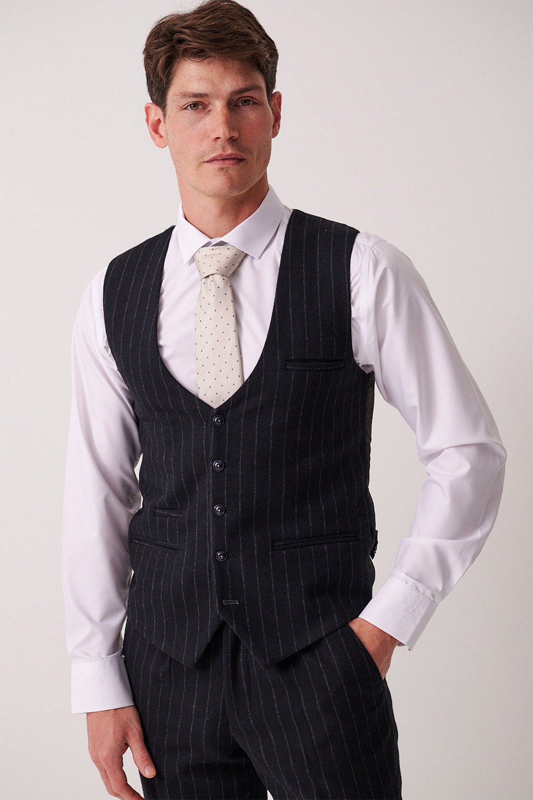 A man wearing the marcdarcy HANSON Navy Tweed Pinstripe Three Piece Suit, paired with a slim fit white shirt and a light dotted tie, stands with one hand in his pocket, looking at the camera against a plain background.