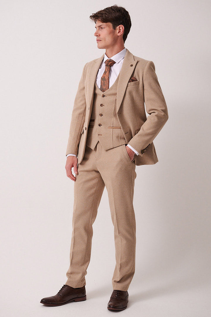 A man stands against a plain background wearing the marcdarcy HAYDEN - Oak Crossweave Three Piece Suit with a matching vest, white shirt, patterned tie, brown pocket square, and brown dress shoes. He looks to the side with one hand in his pocket.