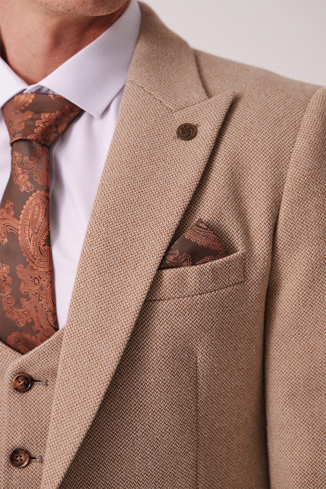 A close-up shows a man in marcdarcy’s HAYDEN - Oak Crossweave Blazer with brown buttons, paired with a white dress shirt and brown paisley tie and pocket square. The blazer’s textured fabric is visible; only his lower face is partially seen.
