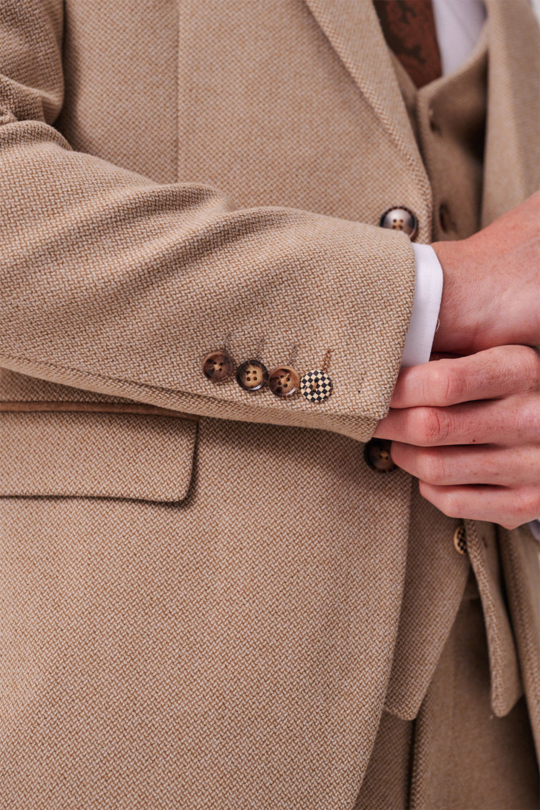 Close-up of a person adjusting the cuff of a marcdarcy HAYDEN - Oak Crossweave Three Piece Suit, featuring brown buttons, a white shirt cuff, and matching vest.