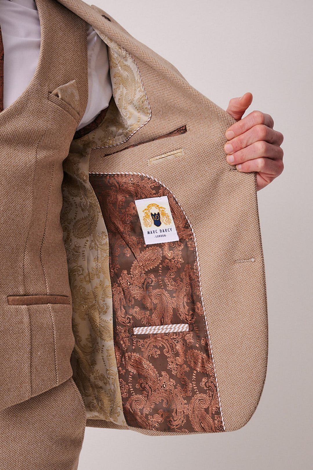 A person holds open a beige marcdarcy HAYDEN - Oak Crossweave Three Piece Suit jacket, revealing a patterned brown and gold lining with an embroidered label. The slim fit suit includes a matching vest and visible pockets.