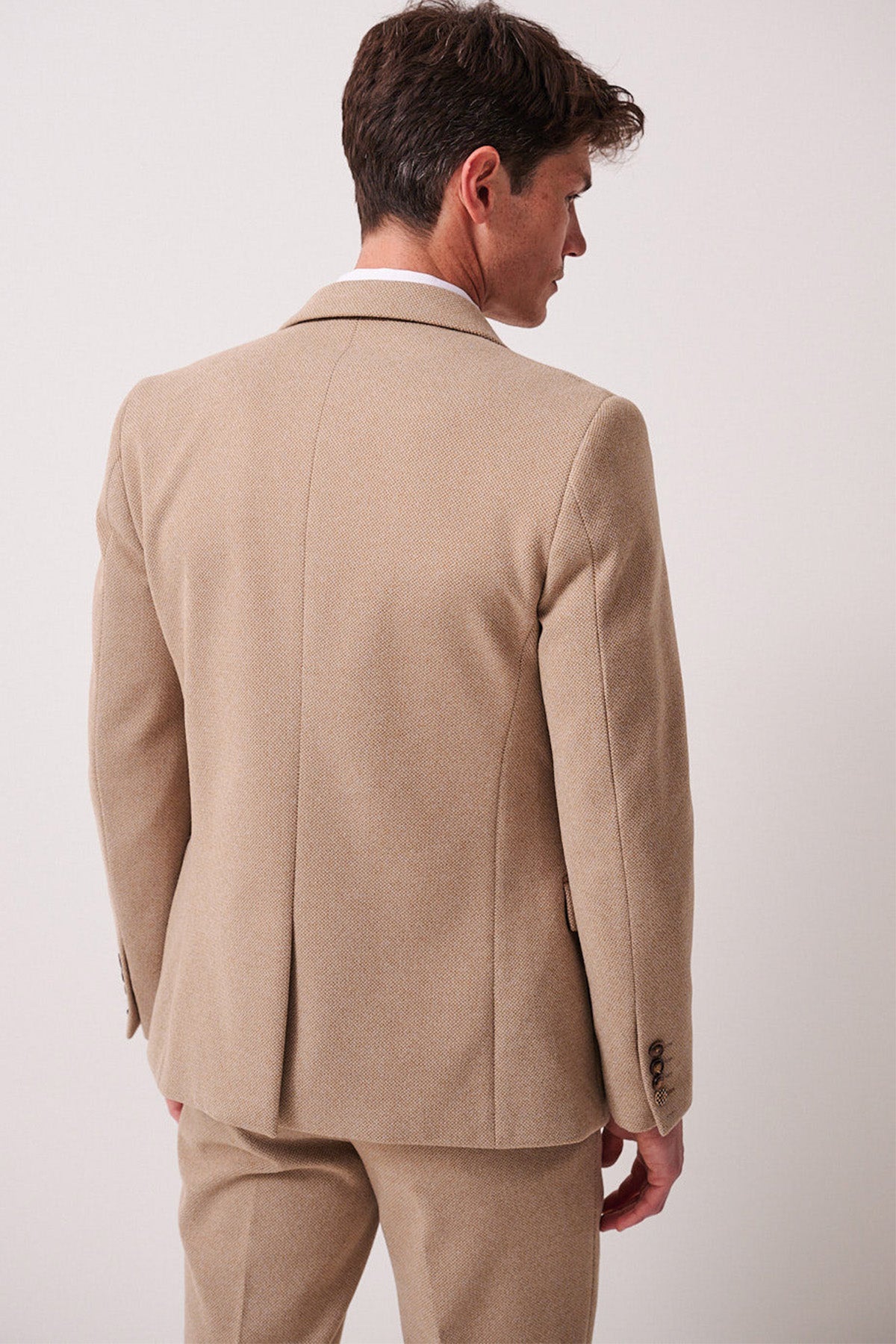 A man wearing the marcdarcy HAYDEN Oak Crossweave Blazer stands with his back to the camera, head turned slightly left, in matching trousers against a plain, light-colored background.