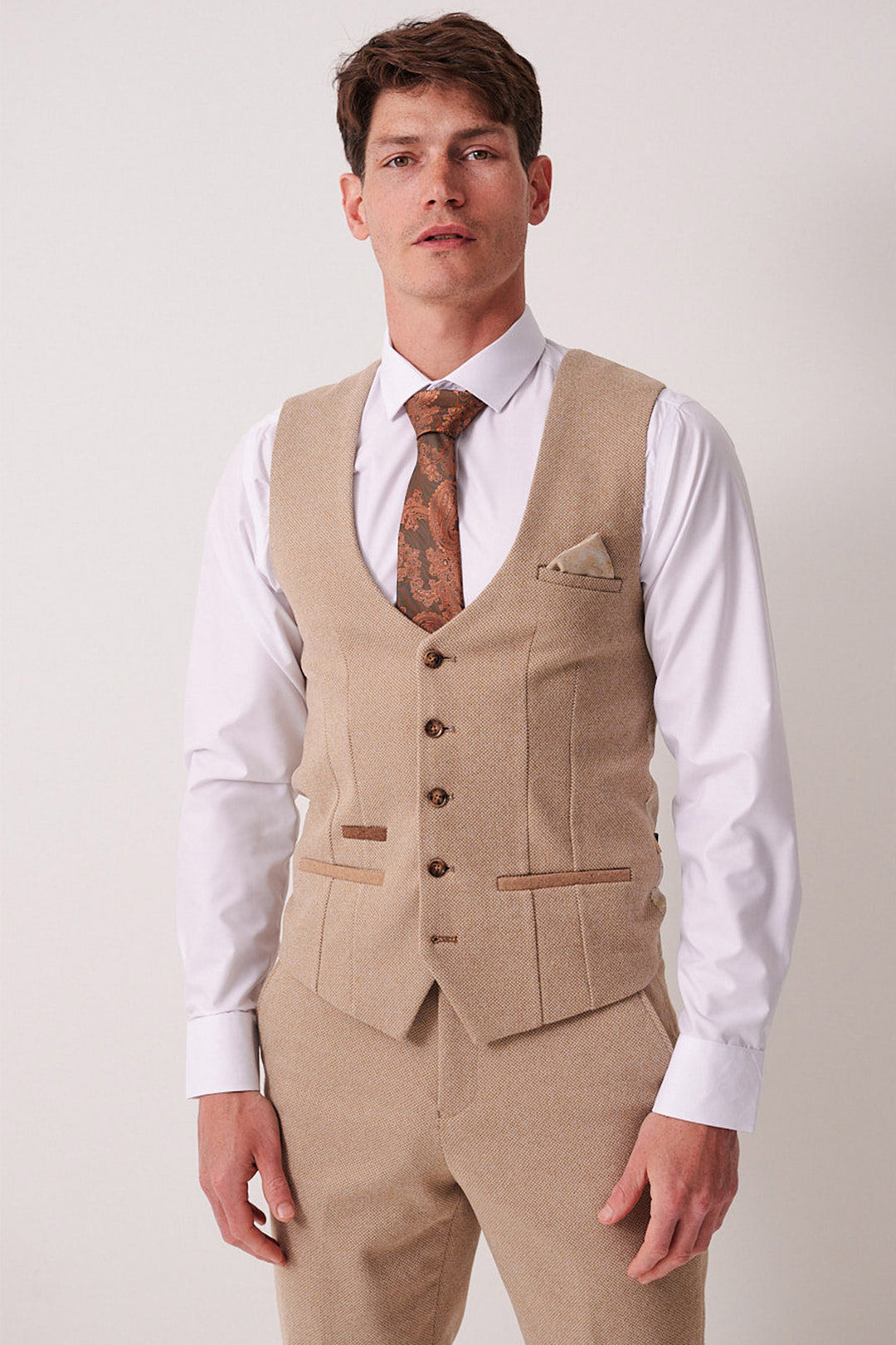 A man wears the Marc Darcy HAYDEN - Oak Crossweave Waistcoat as part of a slim fit beige three-piece suit with a white shirt, brown patterned tie, and matching pocket square, standing against a plain light background.