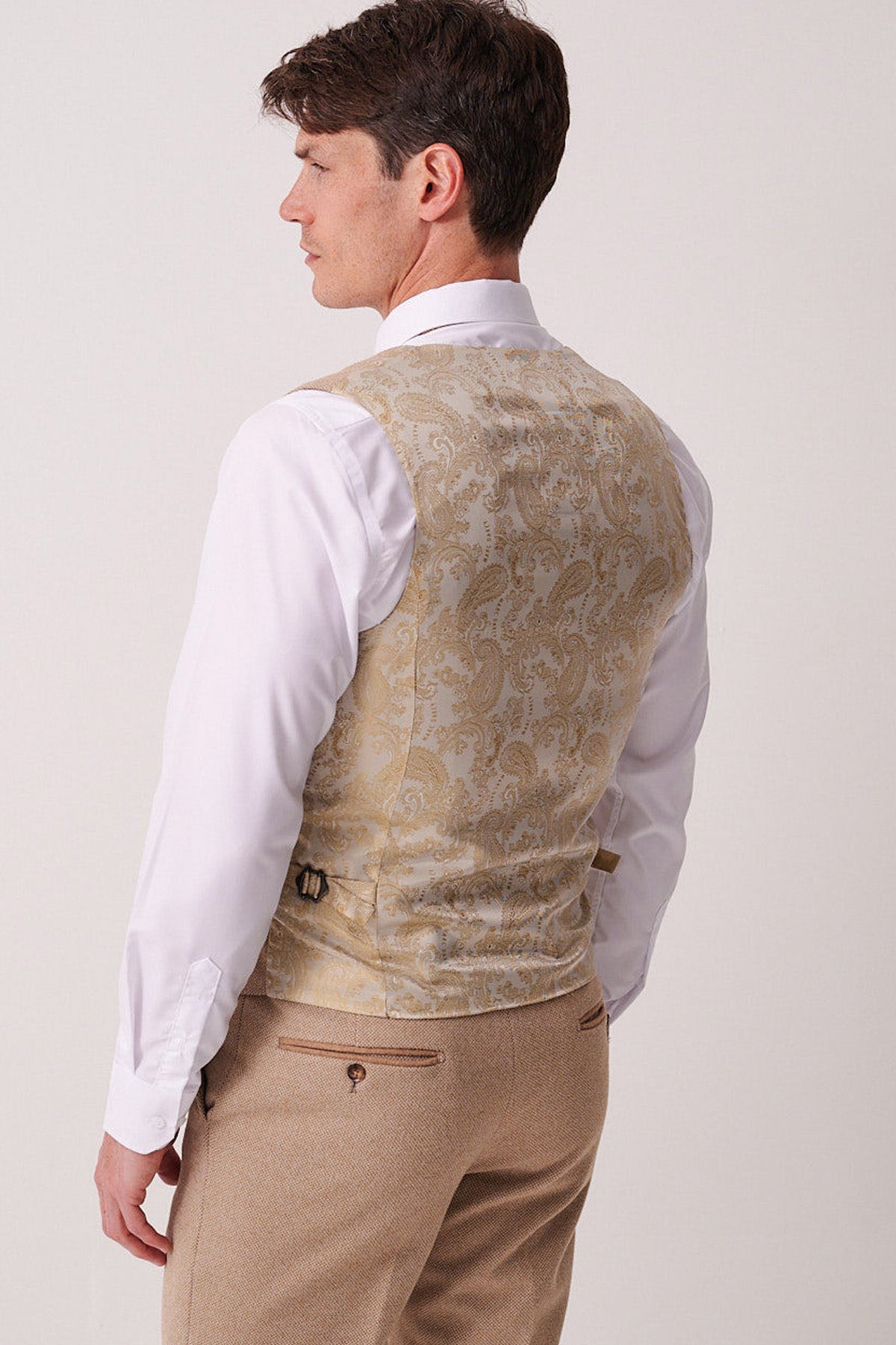 A man faces away, displaying the back of his slim fit white shirt, Marc Darcy HAYDEN Oak Crossweave Waistcoat in light gold with pattern detail, and beige dress pants against a plain light background.