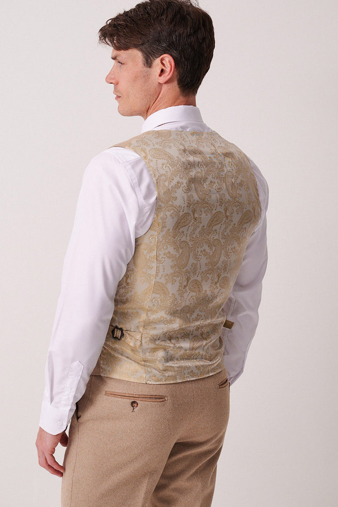 A man models the marcdarcy HAYDEN - Oak Crossweave Three Piece Suit, featuring a white dress shirt, beige paisley vest with adjustable strap, and tan trousers. The plain light background highlights this elegant tweed suit ensemble.