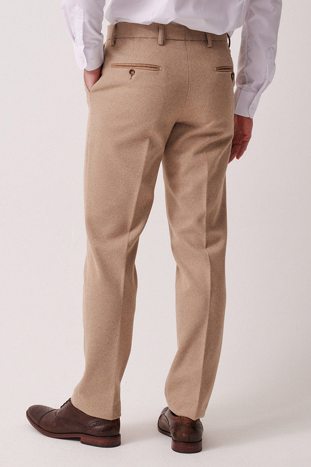 A person wearing marcdarcy HAYDEN - Oak Crossweave Trousers, a white shirt, and brown dress shoes stands facing away with one hand in their pocket against a plain light background.