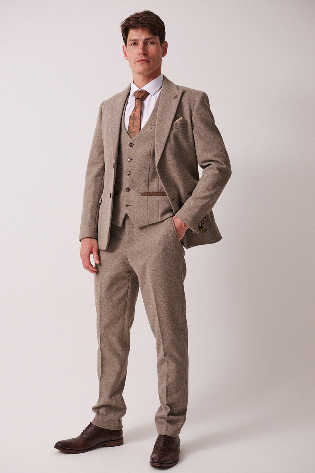 A man models the marcdarcy HAYDEN Tan Crossweave Three Piece Suit with a white shirt, patterned tie, and brown dress shoes. One hand is in his pocket as he looks ahead against a plain background.