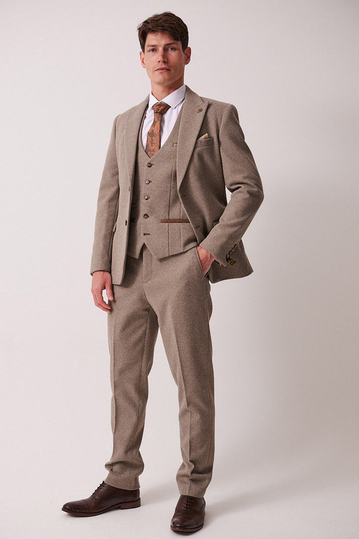 A man models the marcdarcy HAYDEN Tan Crossweave Three Piece Suit with a white shirt, patterned tie, and brown dress shoes. One hand is in his pocket as he looks ahead against a plain background.