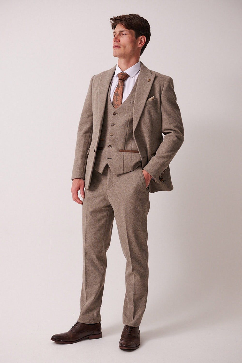 A man in the marcdarcy HAYDEN Tan Crossweave Three Piece Suit with a white shirt and patterned brown tie stands with his hand in his pocket.