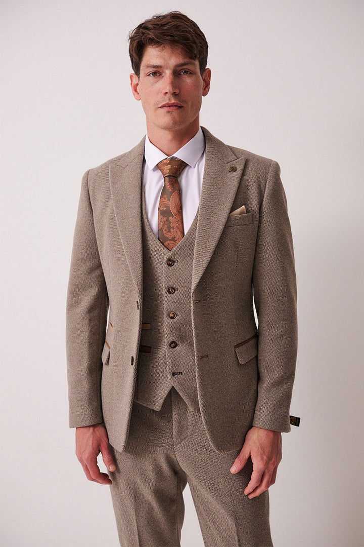 A man stands against a plain light background wearing the marcdarcy HAYDEN Tan Crossweave Three Piece Suit, styled with a white shirt and patterned brown tie. His expression is neutral and his hands rest relaxed at his sides.