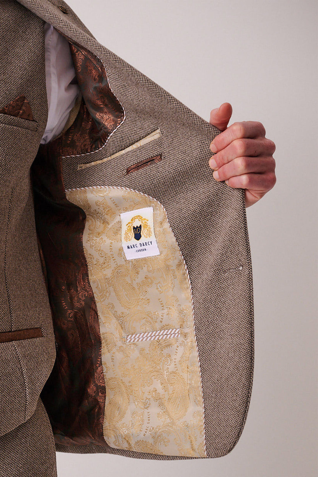 A person holds open the front of a marcdarcy HAYDEN Tan Crossweave Three Piece Suit blazer, showing its ornate golden paisley lining and a label featuring the marcdarcy logo of a man in a hat.
